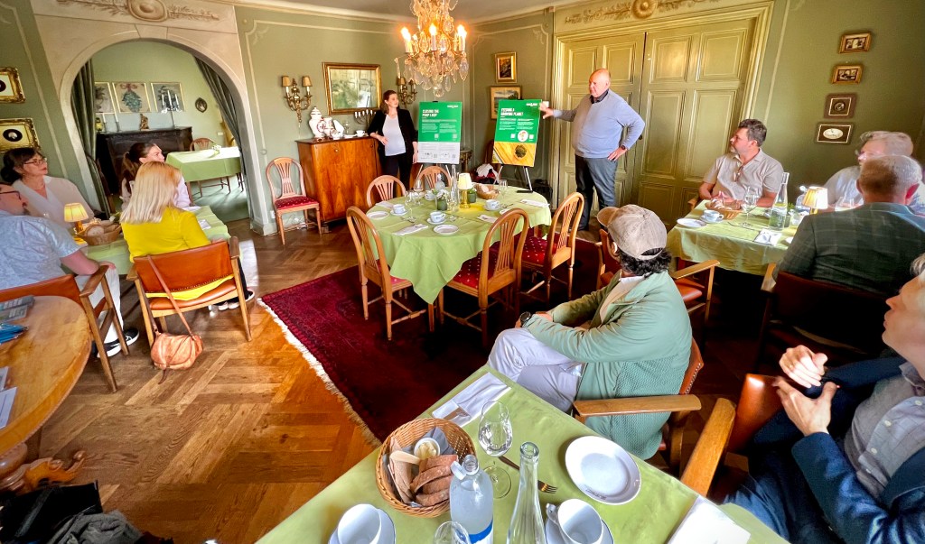 A group of people seated around tables in a dining setting, attentively listening to a speaker presenting in front of a green informational poster.