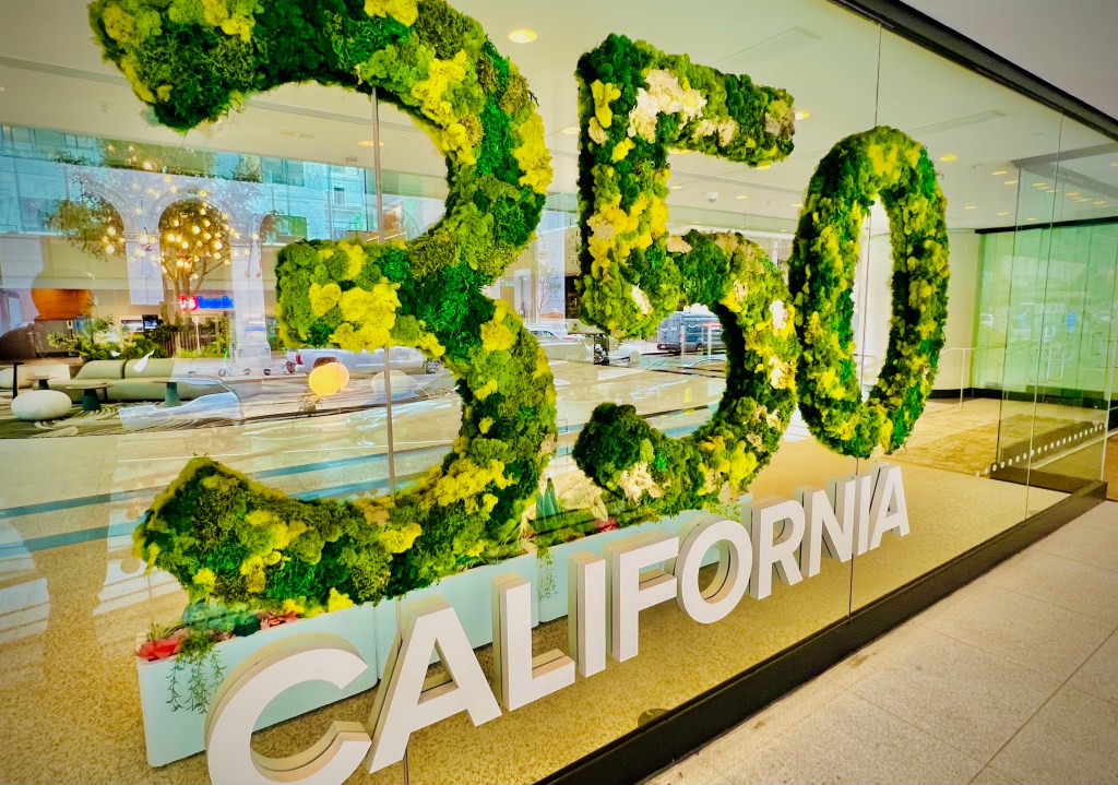 A large decorative display featuring the number '350' and the word 'CALIFORNIA', made from vibrant green and yellow foliage, set against a glass backdrop in a contemporary indoor space.