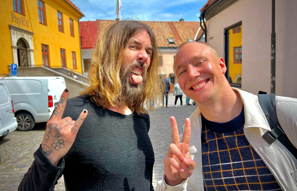 Two men posing for a photo in a street with historic buildings in the background. One man is making a rock sign gesture while the other is showing a peace sign, both smiling.