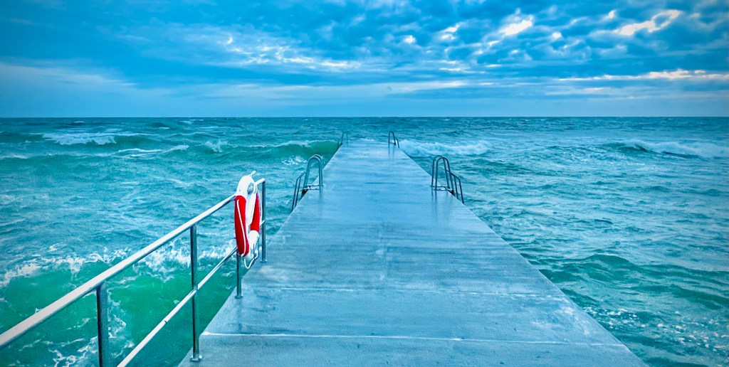 A wide concrete pier extending into the ocean, surrounded by turbulent waves and a cloudy blue sky.