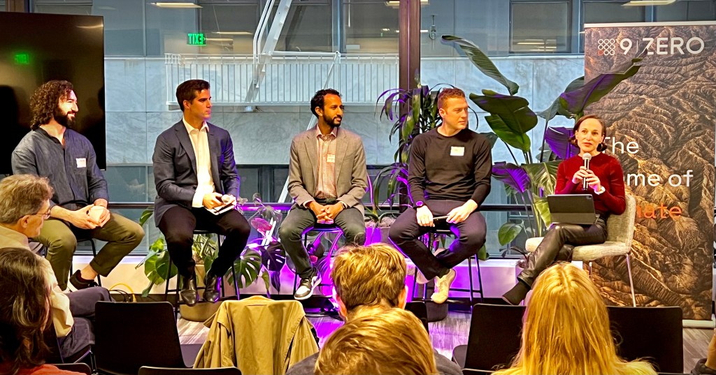 A panel discussion featuring five speakers seated in front of an audience, with plants and a digital display in the background.
