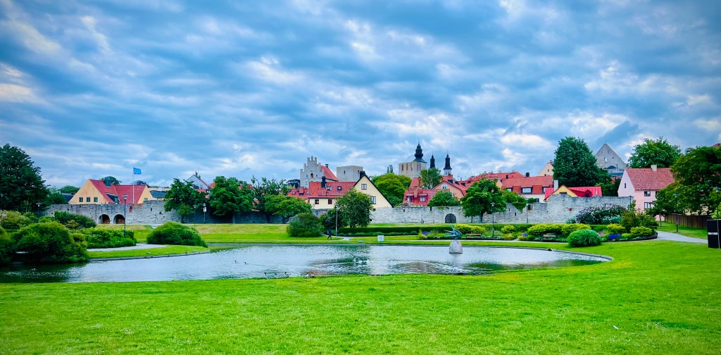 A scenic view of Visby on the island of Gotland, showcasing a lush green park with a pond, surrounded by historic buildings and cloudy skies.