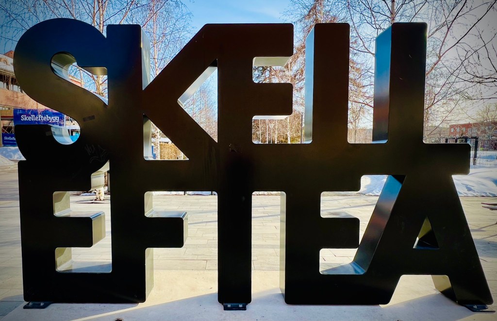 A large black sculpture displaying the word 'SKELLEFTEA' in a bold, stylized font against a clear blue sky.