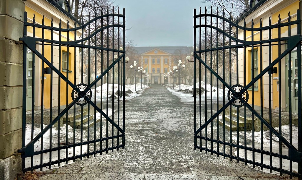 An open black wrought iron gate leading to a snowy pathway lined with lampposts, with a yellow building visible in the background under a foggy sky.