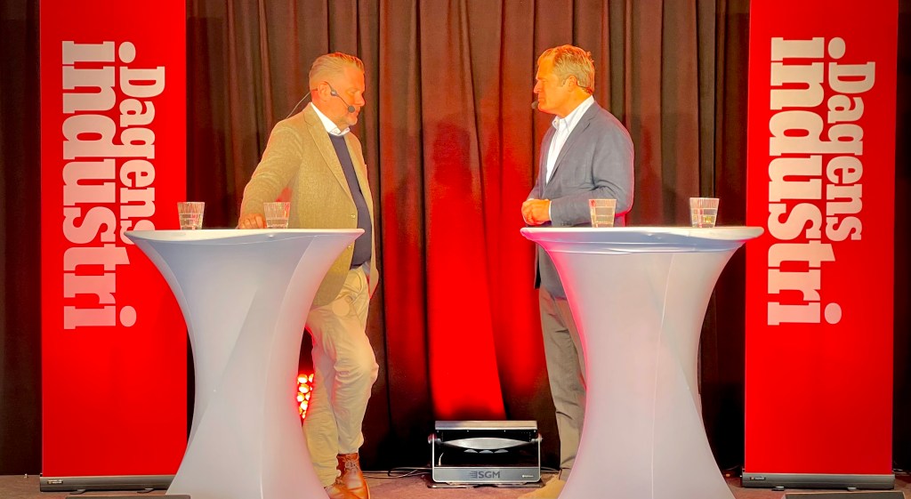 Two speakers engaged in conversation at an event, standing behind white tables with glasses of water, in front of banners with 'Dagens Industri' branding.