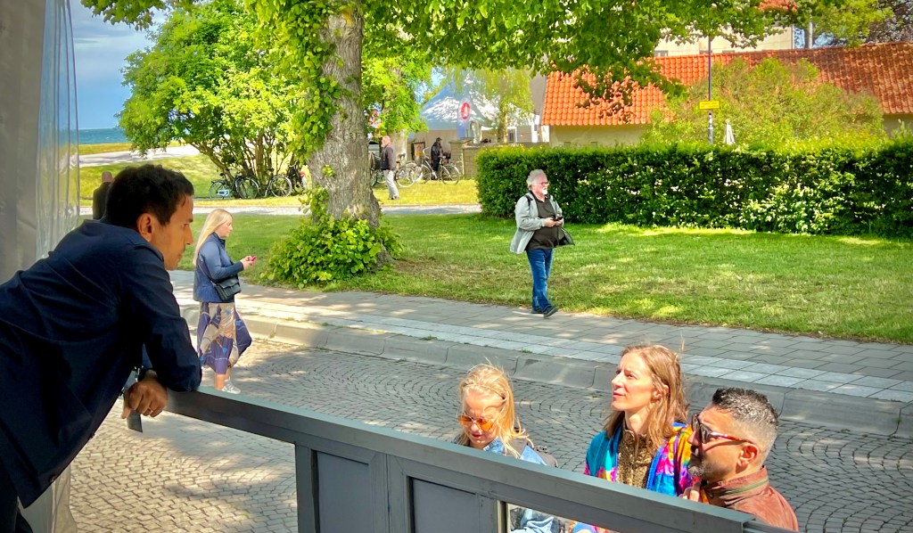 A group of people engaged in conversation outdoors, with a view of trees and buildings in the background. One person leans over a railing while others listen attentively. In the background, pedestrians walk along a path.