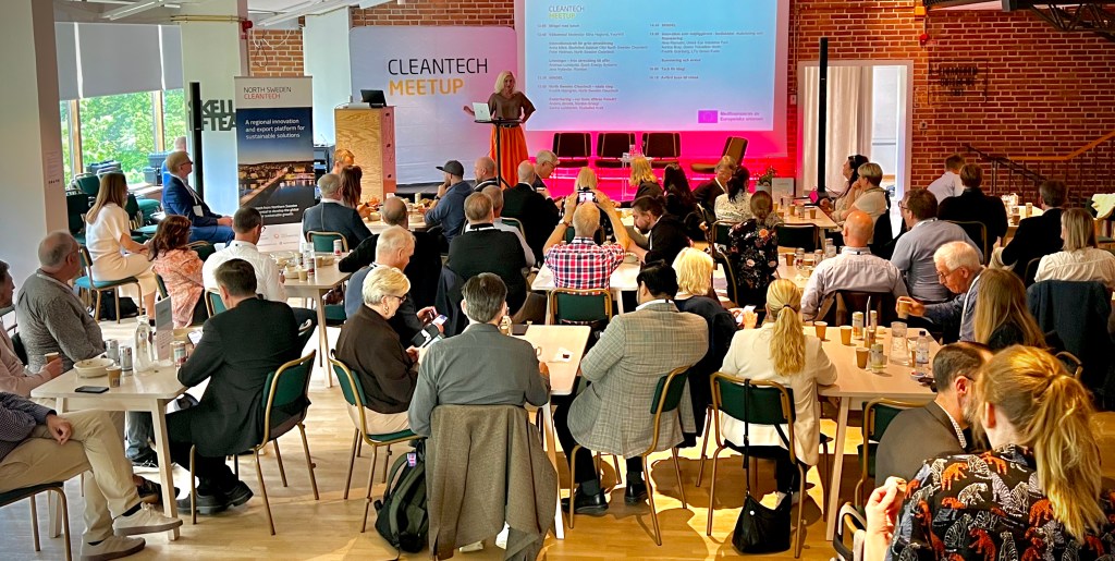 Attendees at a Cleantech Meetup event sitting at tables, with a speaker presenting in front of a large screen displaying the agenda.