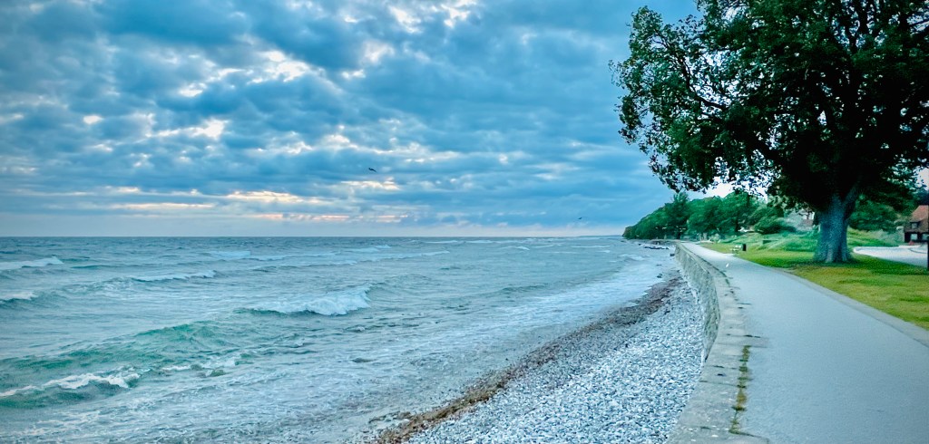 A serene view of the coastline in Visby on Gotland, featuring calm waves lapping against a pebbled shore with a tree-lined pathway and cloudy skies in the background.